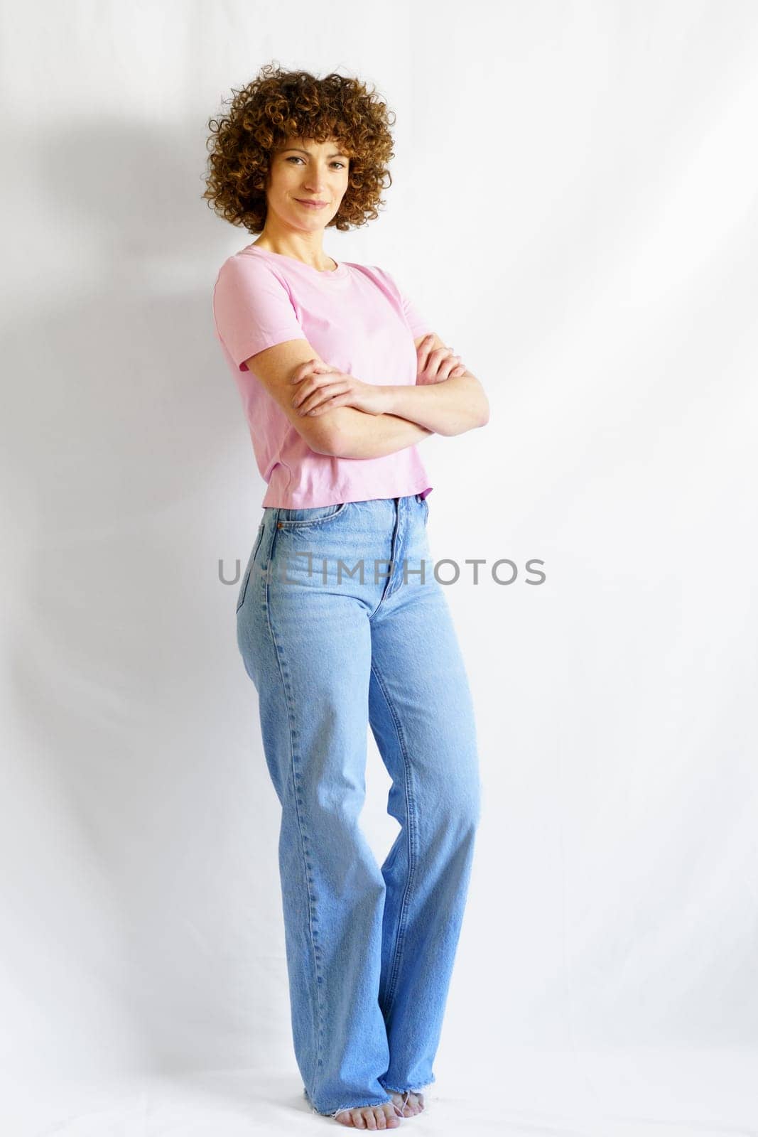 Full body of curly haired woman in casual clothes leaning on white wall while standing with hands crossed looking at camera and smiling