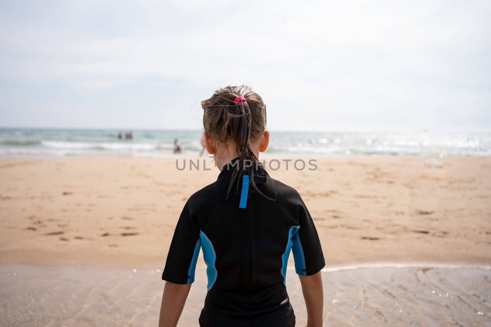 Little girl surfer in wetsuit standing ocean beach. Child after surfing on sandy beach back view