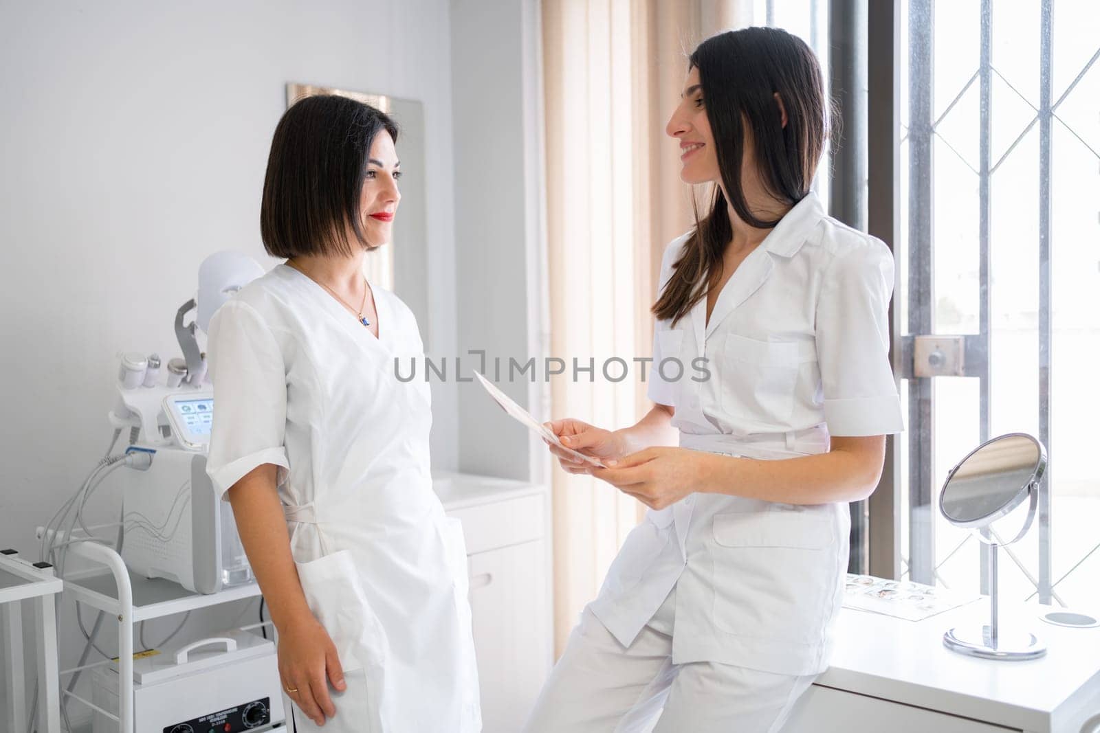 Two female doctors standing in medical clinic, discussing results of patient's exams. looking at each other and smiling. Healthcare specialists are having a professional conversation.