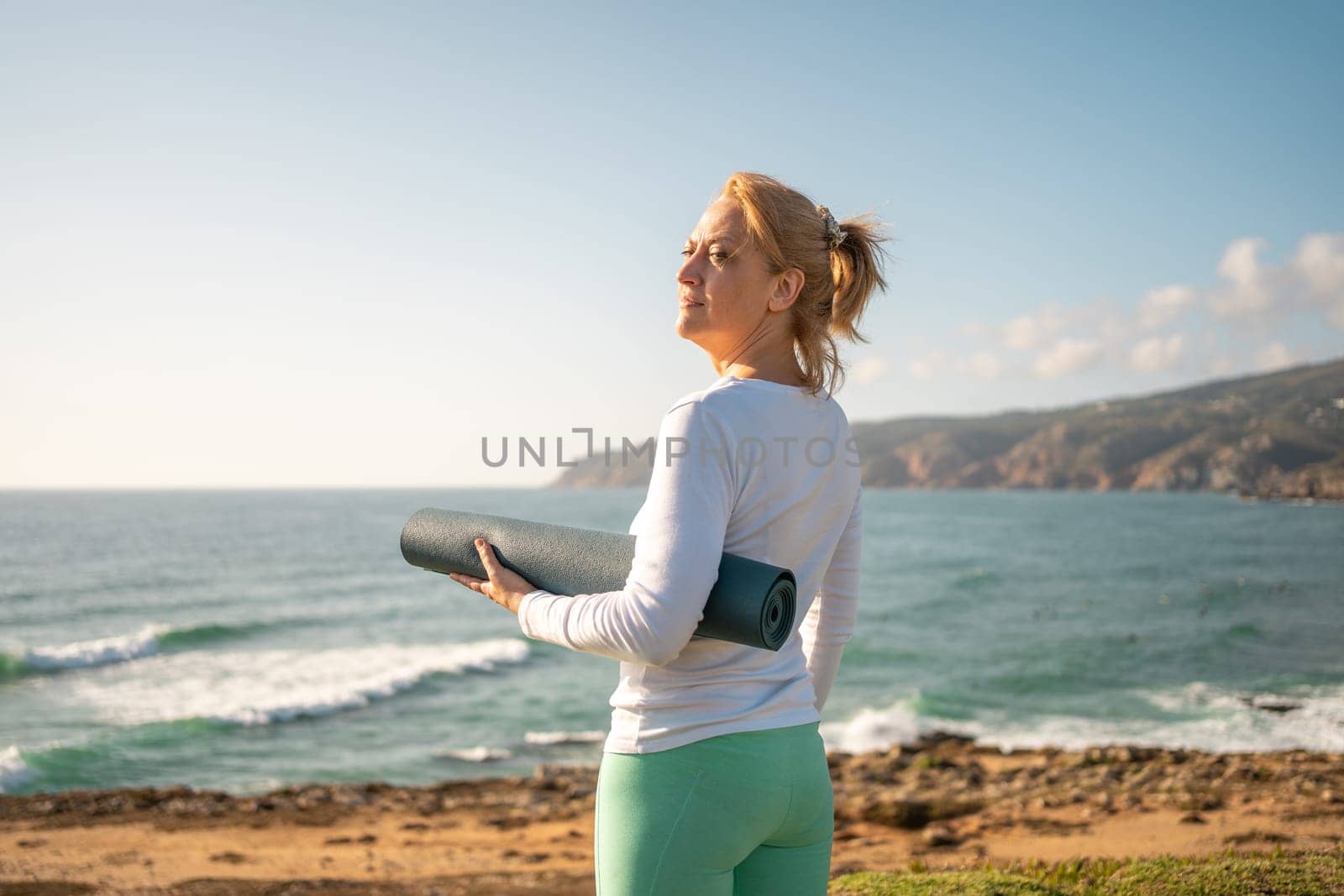Senior woman standing ocean shore, holding yoga mat. She appears to be happy and enjoying the scenic views before her yoga session. This depicts a harmonious and healthy senior lifestyle concept.