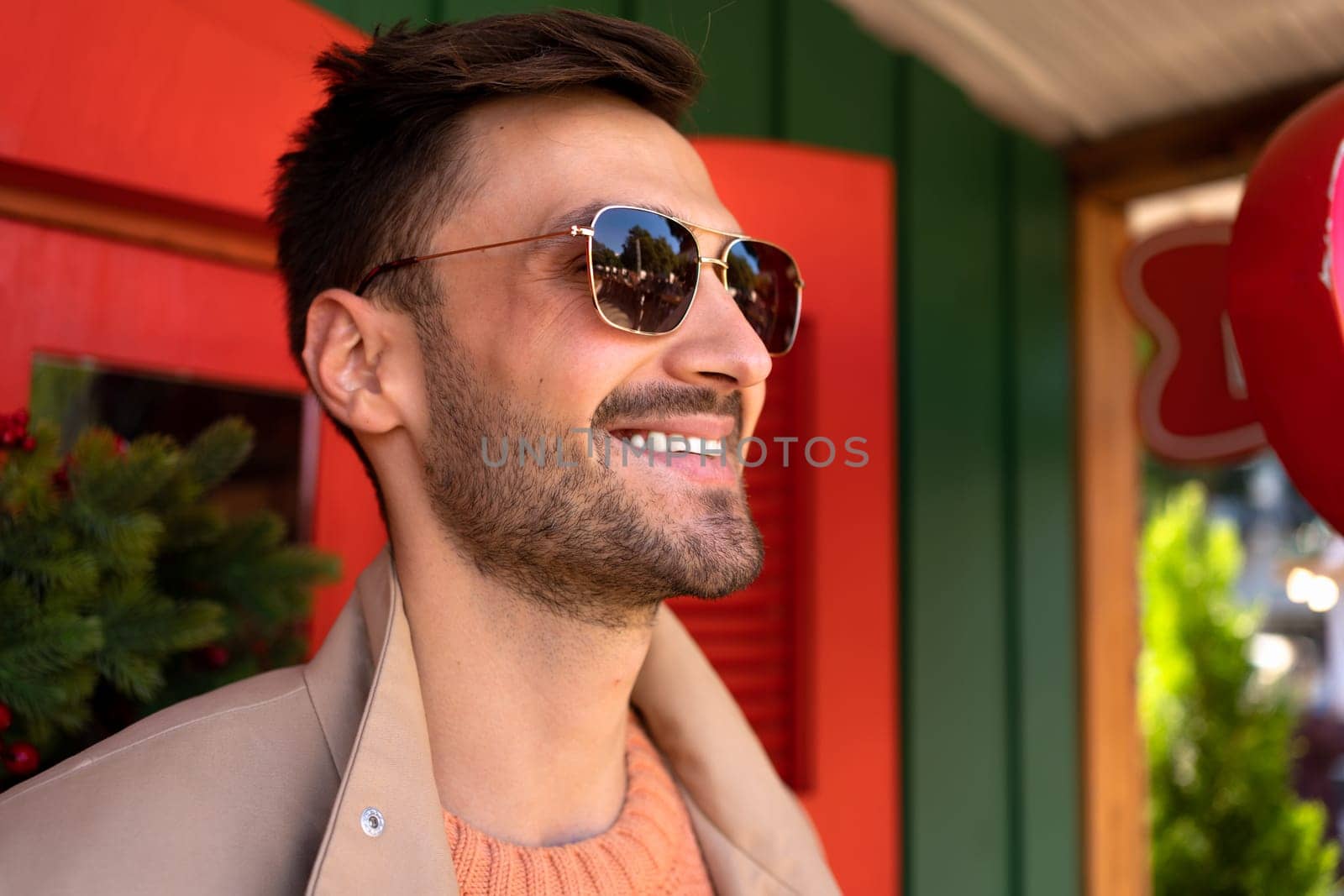 Handsome man wearing sunglasses and trench coat standing outside looking forward and smiling. Close up portrait young bearded male person