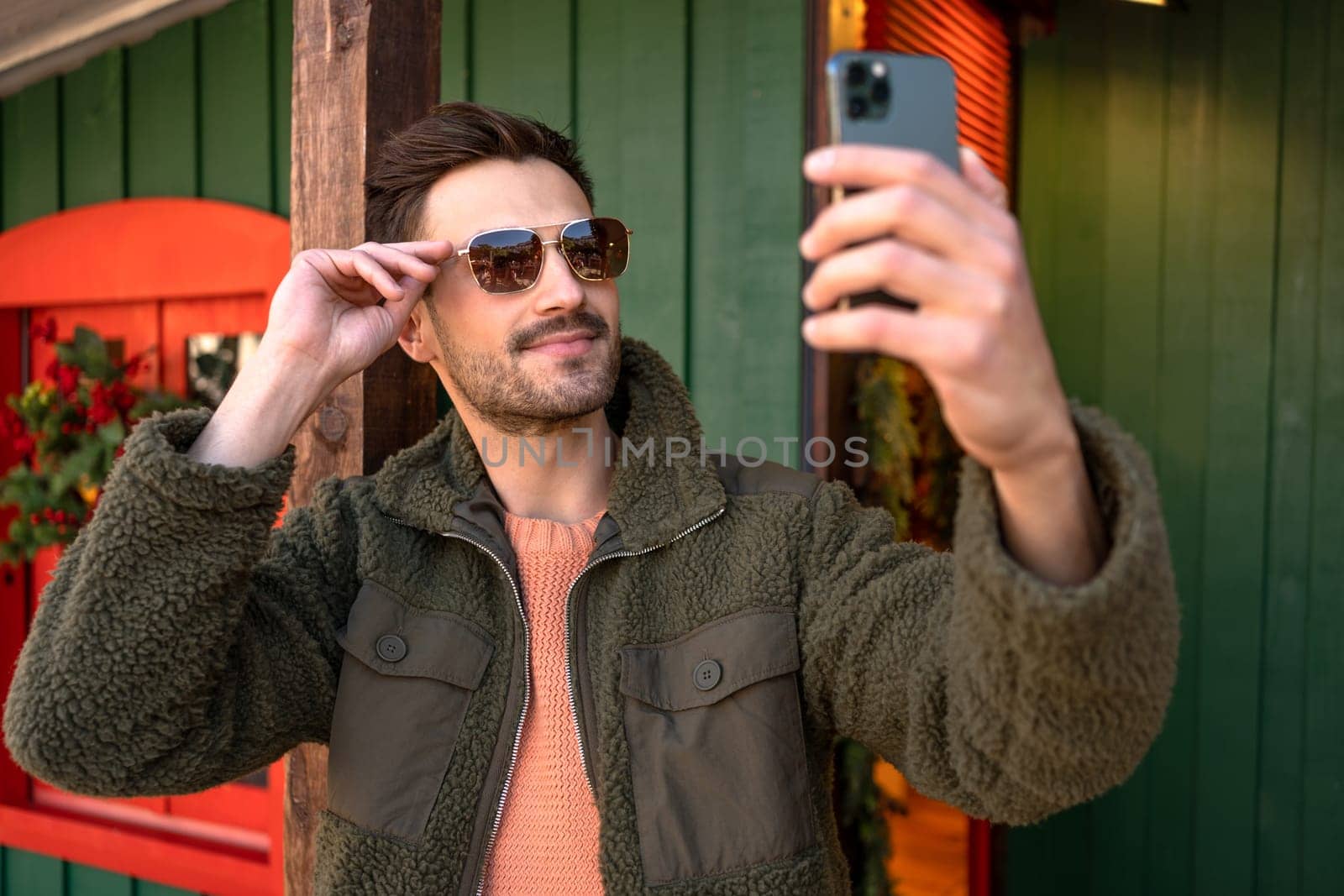 Handsome young man taking self portrait while walking through the city. Urban millenial with beard wearing sunglasses using smartphone and smiling carefree outdoors. Lifestyle concept.