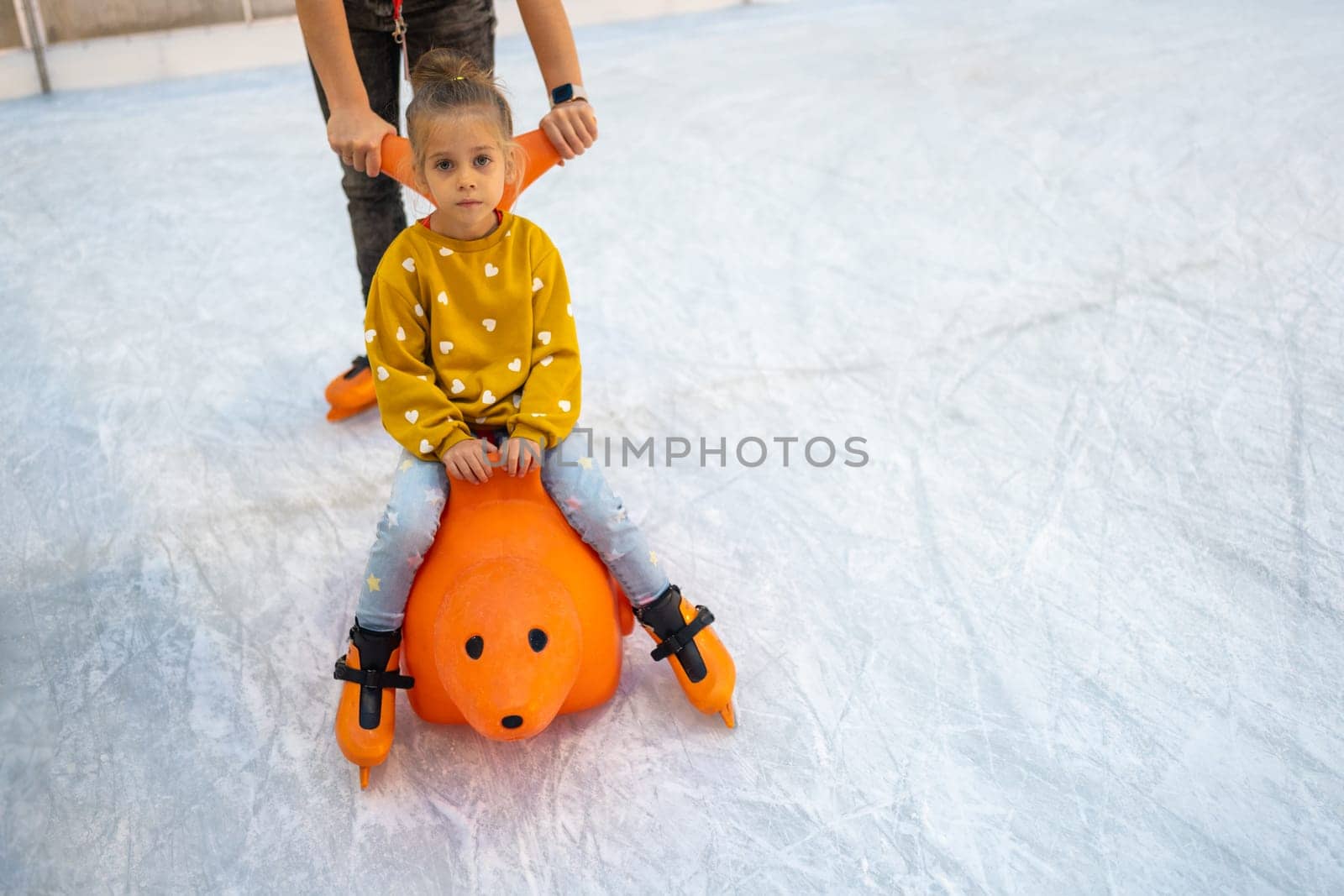 Mom with daughter have fun on ice skater rink use skating aid dolphin. Mom teaches little daughter to skate. Winter Christmas ice rink