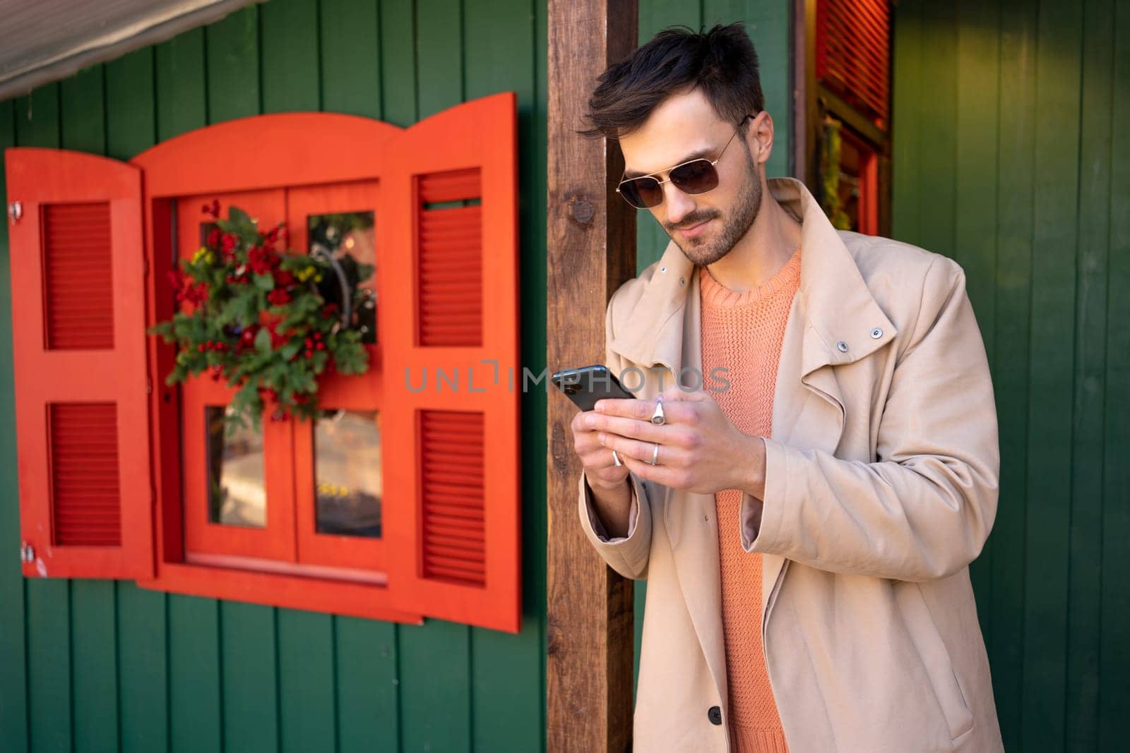 Stylish man wearing sunglasses and trench coat standing outside on Christmas market in European city. Using his smartphone to send holiday greetings enjoying festive atmosphere