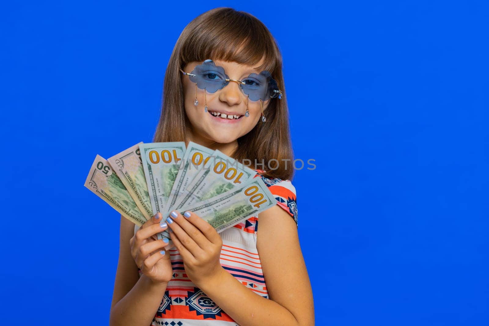 Rich pleased young cute school girl waving money dollar cash banknotes bills like a fan, success, lottery winner, big income, wealth. Teen brunette female child kid on studio blue background