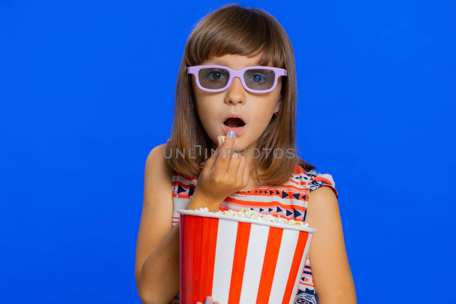 Excited young cute school girl in 3D glasses eating popcorn, watching interesting tv serial, sport game, film, online social media movie content. Teen female child kid on studio blue background