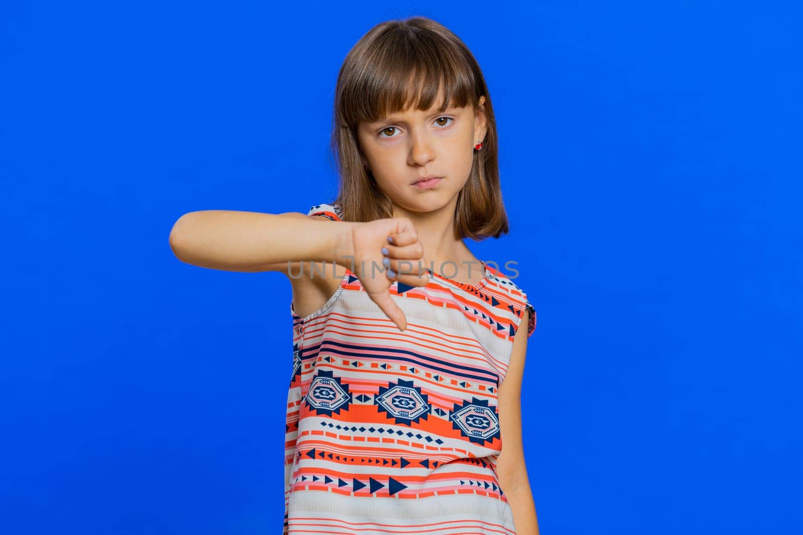 Dislike. Upset young preteen child girl kid showing thumbs down sign gesture, expressing discontent, disapproval, dissatisfied bad work indoors. Displeased cute children on blue studio background