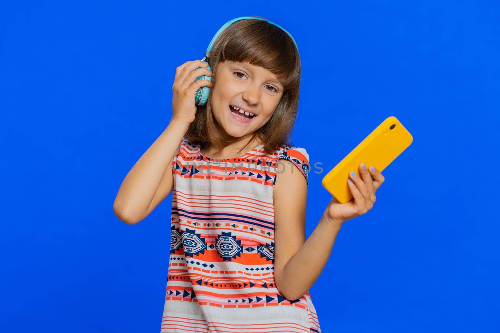 Young school girl listening music via headphones and dancing disco fooling around having fun expressive gesticulating hands. Preteen Caucasian child kid use smartphone app isolated on blue background
