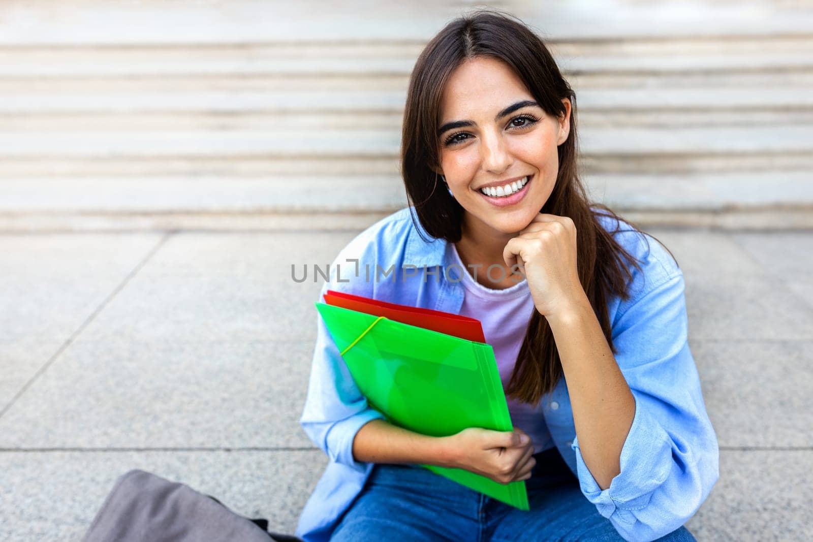 Young beautiful female college student sitting on stairs outside university building looking at camera. Copy space. Education concept.