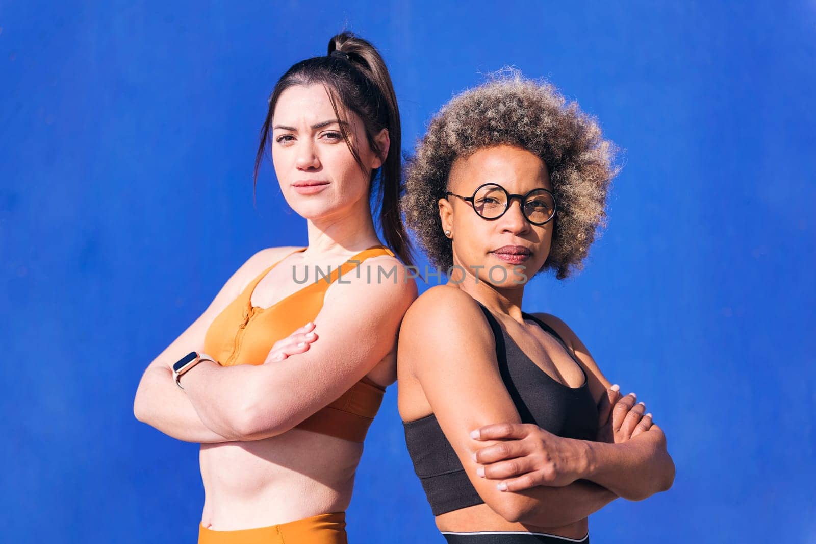 portrait of two proud sportswomen looking at camera with arms crossed in a blue background, concept of real people and sporty lifestyle, copy space for text