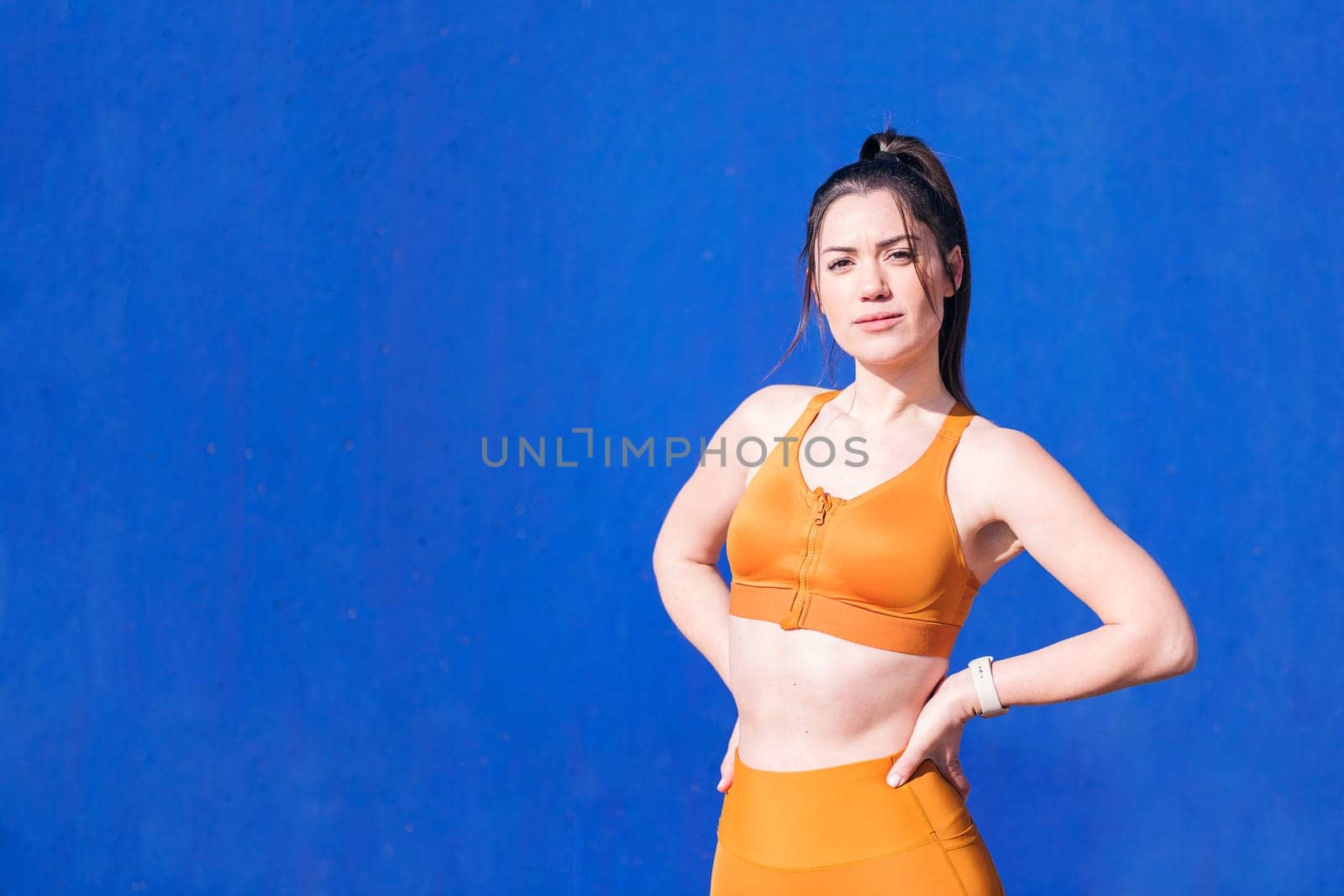 confident caucasian sportswoman with hands on hips in a blue background, concept of real people and sporty lifestyle, copy space for text