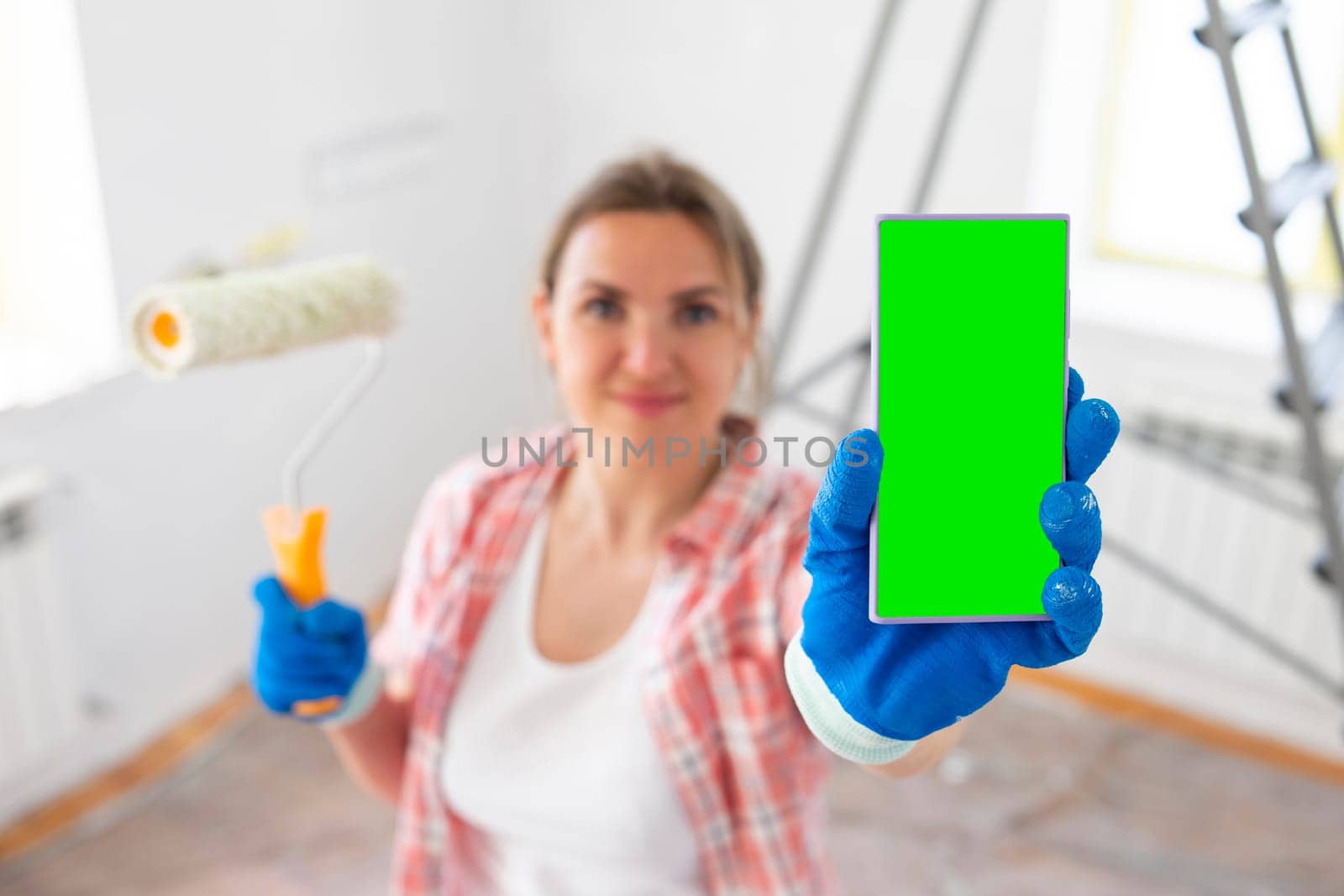 Beautiful woman painter with paint roller showing smartphone with blank screen. Repair concept.