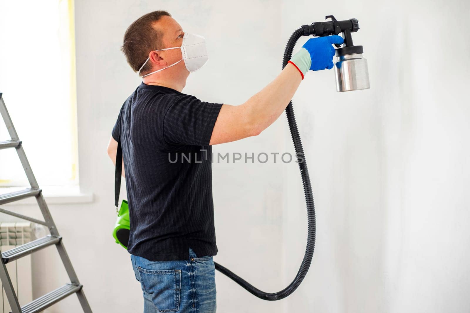 Worker in protective mask painting wall with spray gun. Painting the wall of apartment. Repair concept