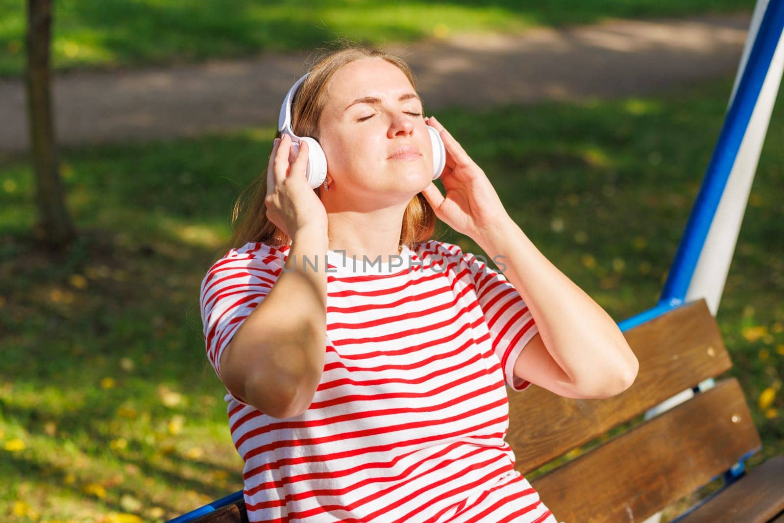 A woman wearing headphones listening to music, breathing fresh air, relaxing while sitting on a bench in sunny day in the park
