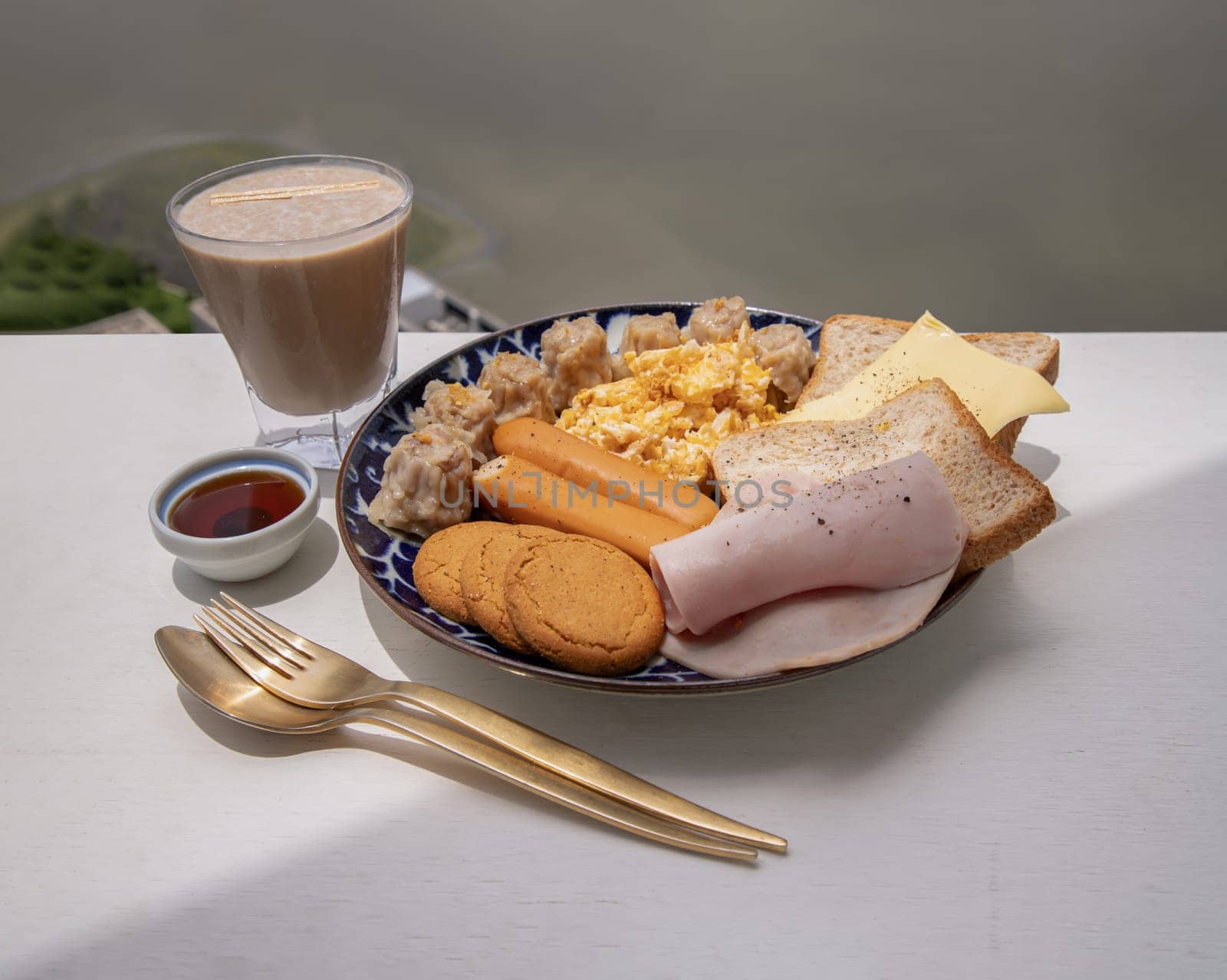 Homemade breakfast is including Breads, Fried sausages, Ham, Pork shumais, Scrambled eggs, Cheese, Crispy ginger cookies and Honey served with Cup of Healthy Protein Shake with River view background. Healthy food concept, Space for text, Selective Focus.
