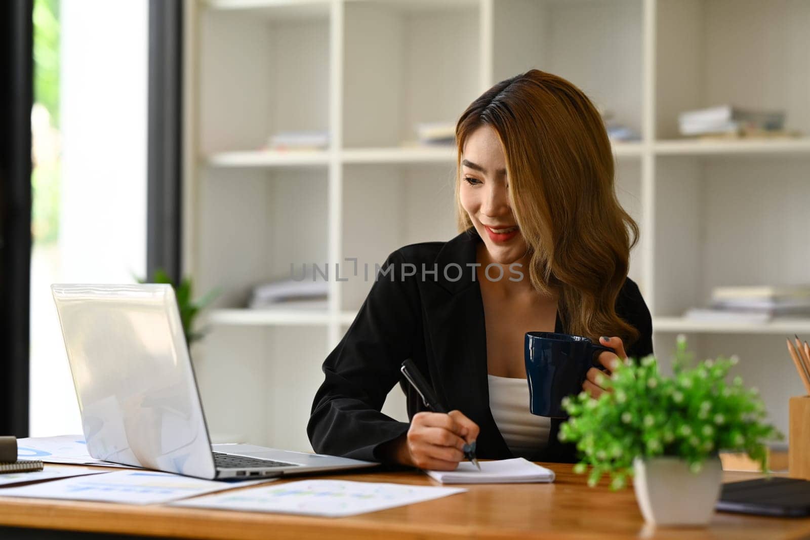 Millennial businesswoman in black suit using laptop, preparing economic report or presentation at workstation by prathanchorruangsak