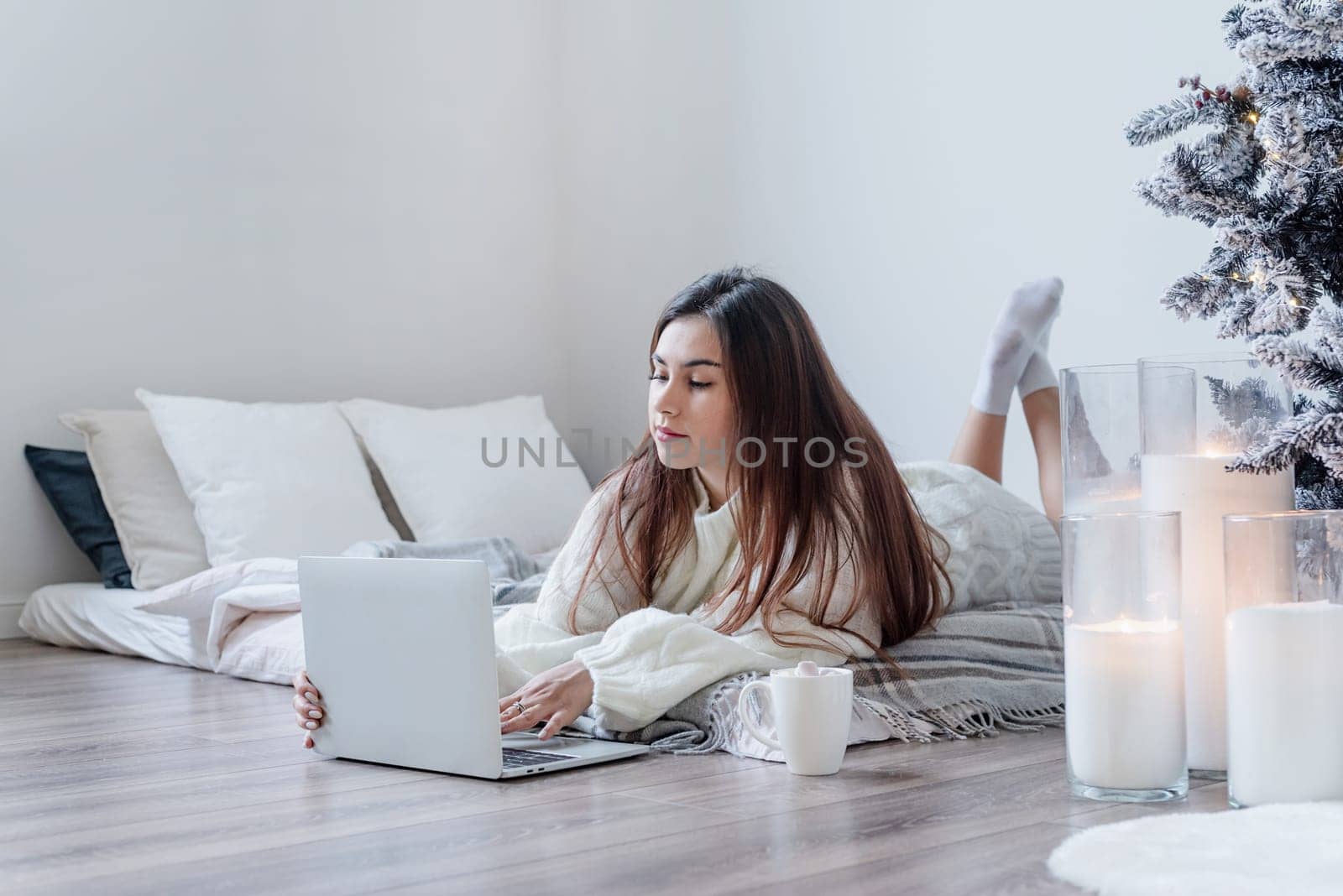 Merry Christmas and Happy New Year. Woman in warm white winter sweater lying in bed at home using laptop, watching movies or working. Christmas tree and candles