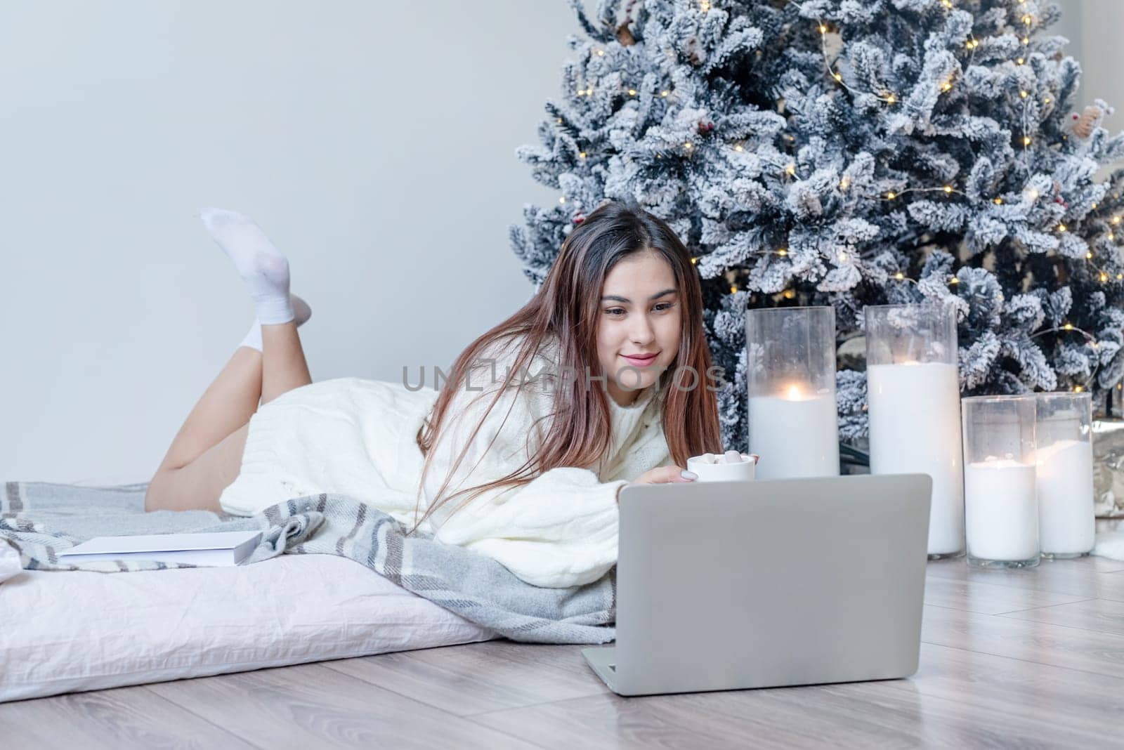 Merry Christmas and Happy New Year. Woman in warm white winter sweater lying in bed at home using laptop, watching movies or working. Christmas tree and candles