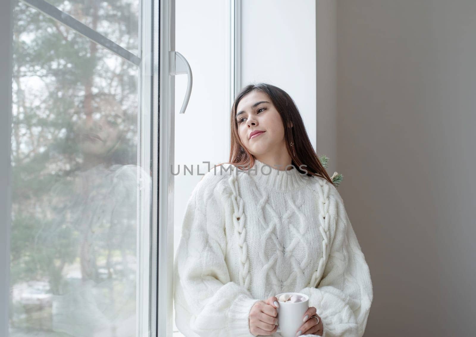 Woman in warm white winter sweater standing next to the window at home at christmas eve holding cup with marshmallows, fir tree behind by Desperada