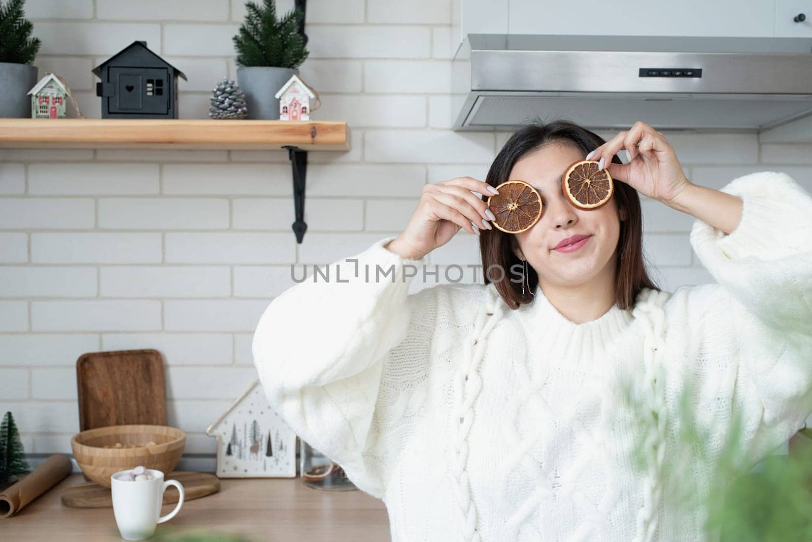 Merry Christmas and Happy New Year. Woman in warm white winter sweater standing at the kitchen at home with dry lemon slices next to face