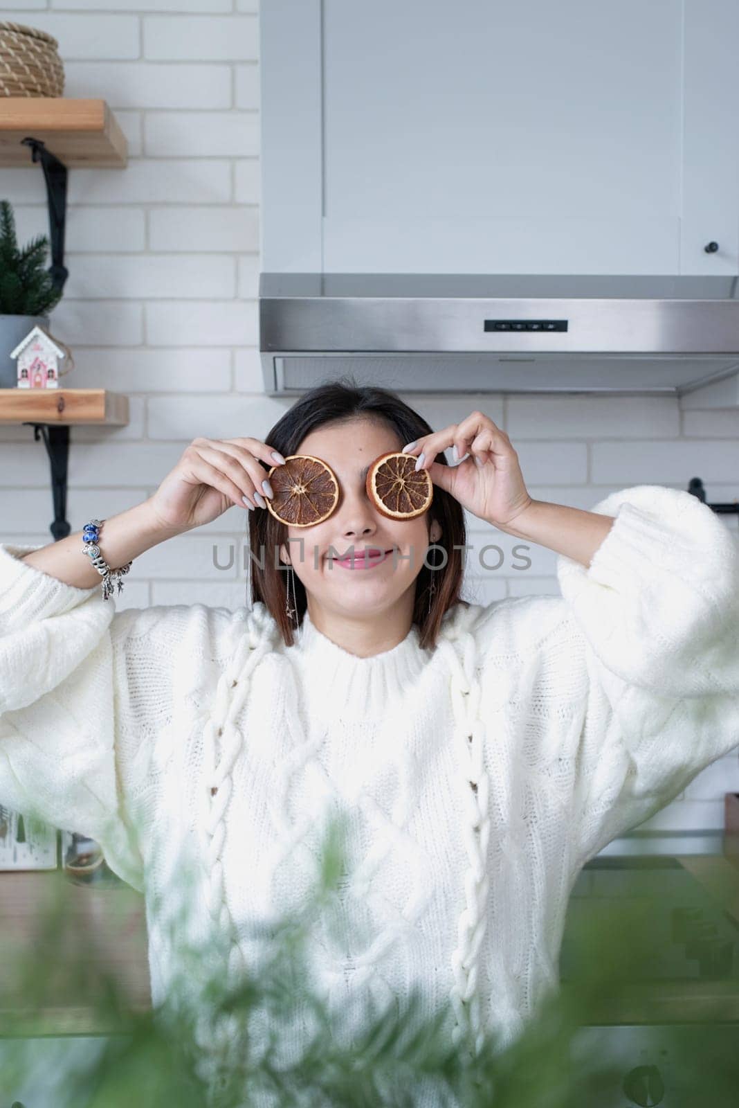 Merry Christmas and Happy New Year. Woman in warm white winter sweater standing at the kitchen at home with dry lemon slices next to face