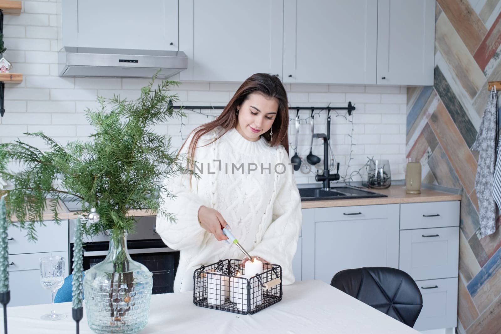 Woman in warm white winter sweater standing at the kitchen at home lighting up the candles by Desperada