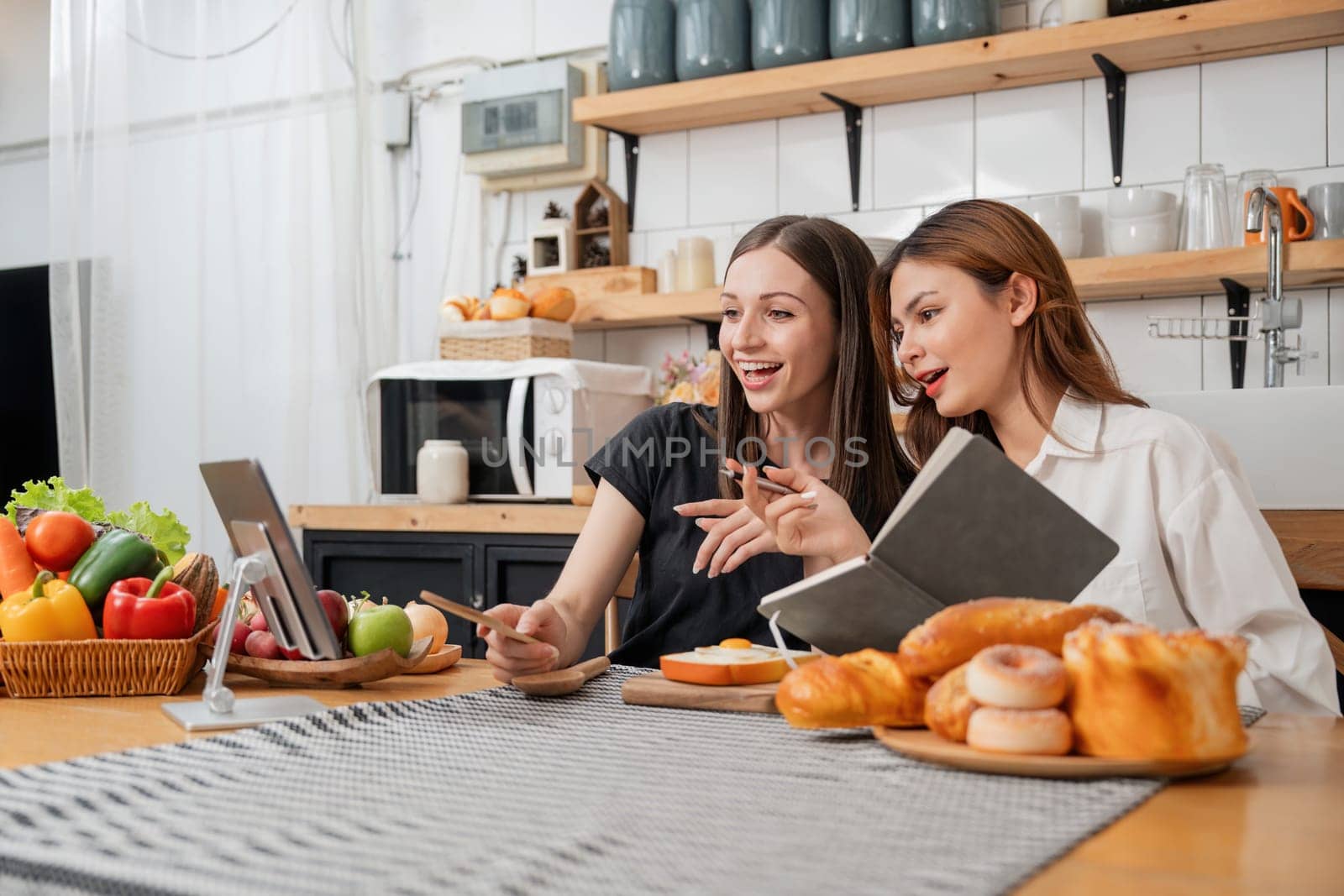 Happy two young women looking laptop computer during cooking together in kitchen room at home. Two young diverse lesbian women spending time together. LGBT and gender identity concept by nateemee