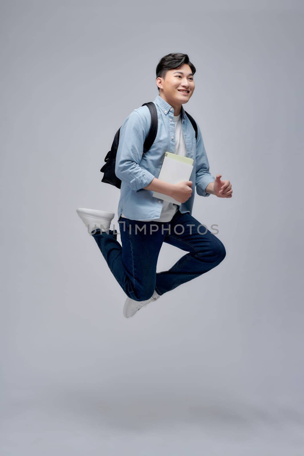 Full length portrait of a smiling young man dressed carrying backpack isolated, holding books, jumping by makidotvn