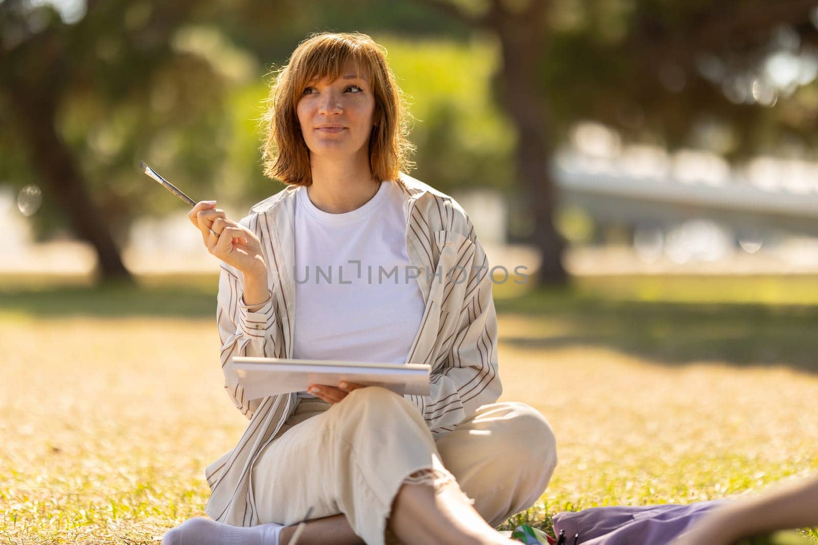 Adult pretty woman sitting in the blooming park holding a painting brush. Mid shot