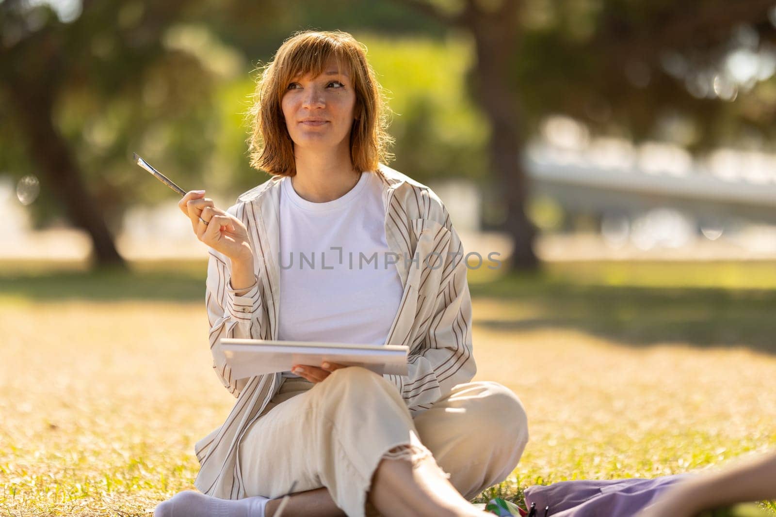 Adult pretty woman sitting in the blooming park holding a painting brush by Studia72