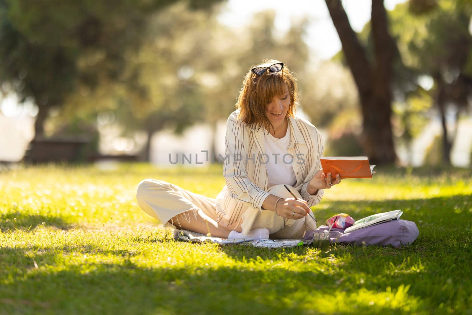 Adult smiling woman painting in her notebook in the blooming park by Studia72