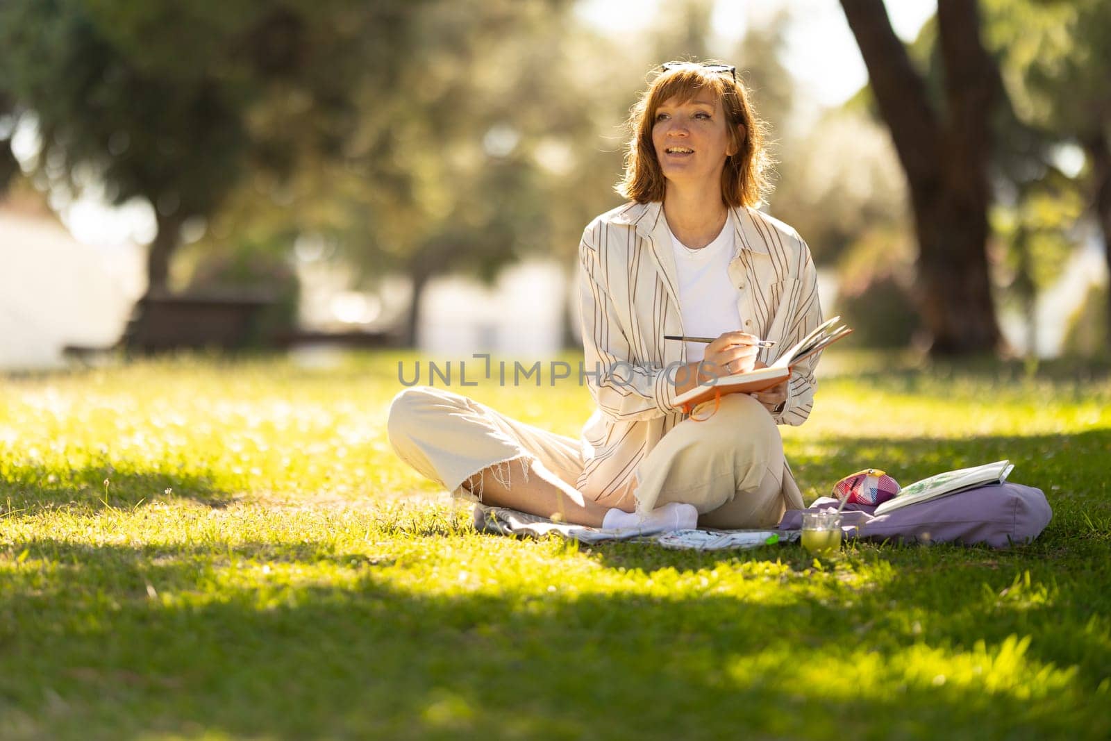 Adult smiling woman painting in her notebook in the blooming park - looking in the distance to the reference. Mid shot