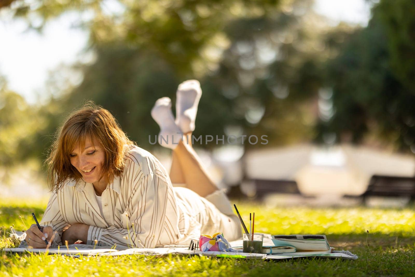 An adult smiling woman lying on the blanket in the green sunny park and drawing in the sketchbook. Mid shot