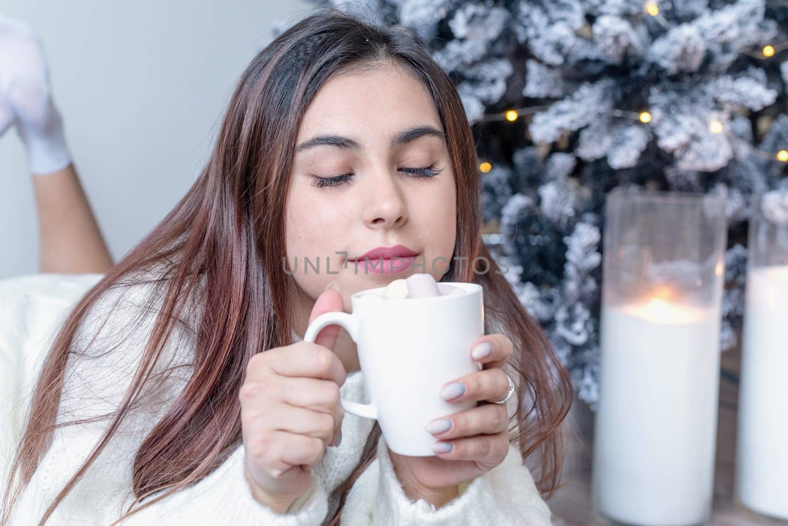 Merry Christmas and Happy New Year. Woman in warm white winter sweater lying in bed at home at christmas eve holding cup with marshmallows, fir tree behind