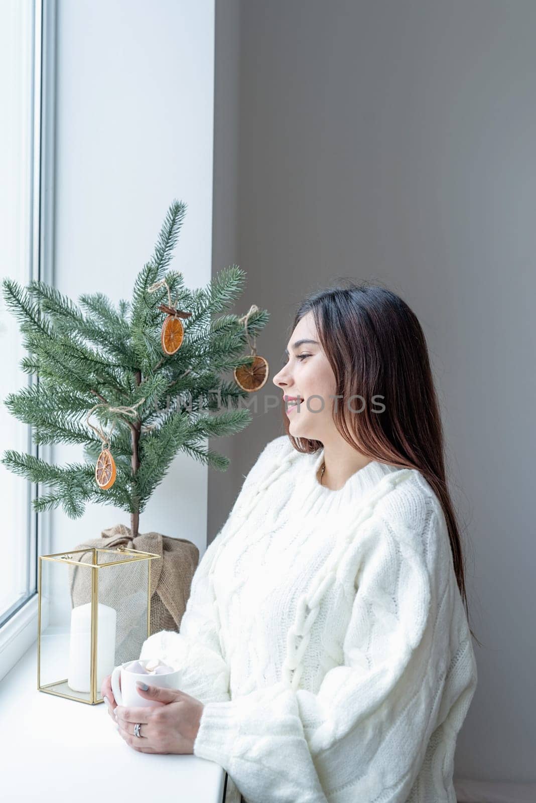 Merry Christmas and Happy New Year. Woman in warm white winter sweater standing next to the window at home at christmas eve holding cup with marshmallows, fir tree behind