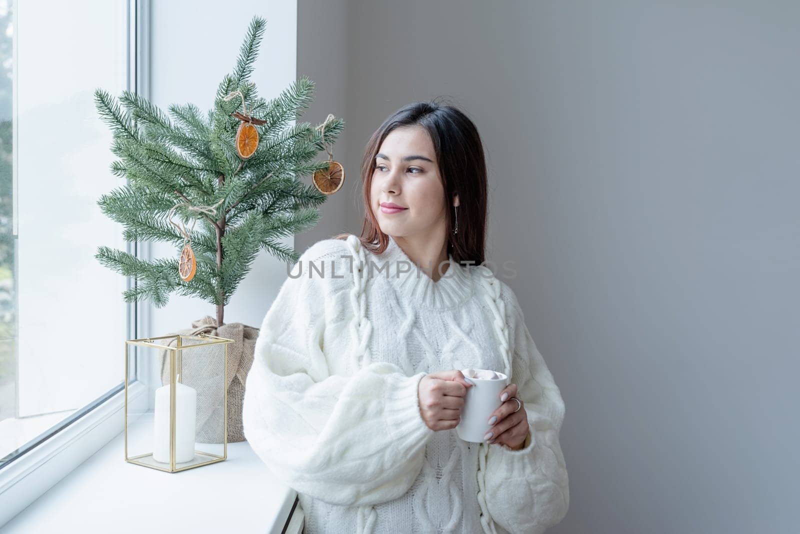 Woman in warm white winter sweater standing next to the window at home at christmas eve holding cup with marshmallows, fir tree behind by Desperada