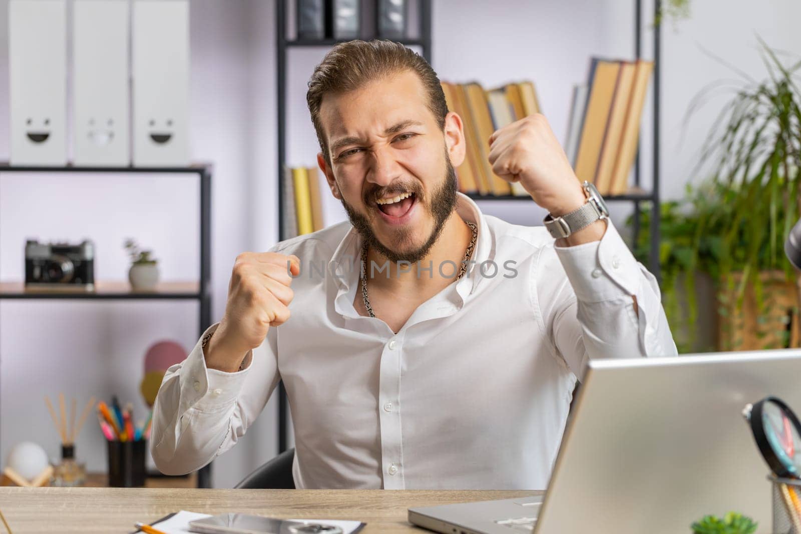 Happy amazed young middle eastern businessman working on laptop shocked by sudden victory, game winning, lottery goal achievemen, good career news. Excited man freelancer at home office workplace desk