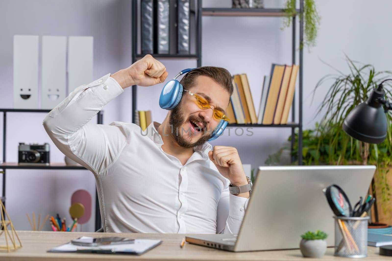 Happy relaxed overjoyed middle eastern businessman working on laptop computer at office wearing headphones listening favorite energetic disco music and dancing. Freelancer man relaxing, taking a break