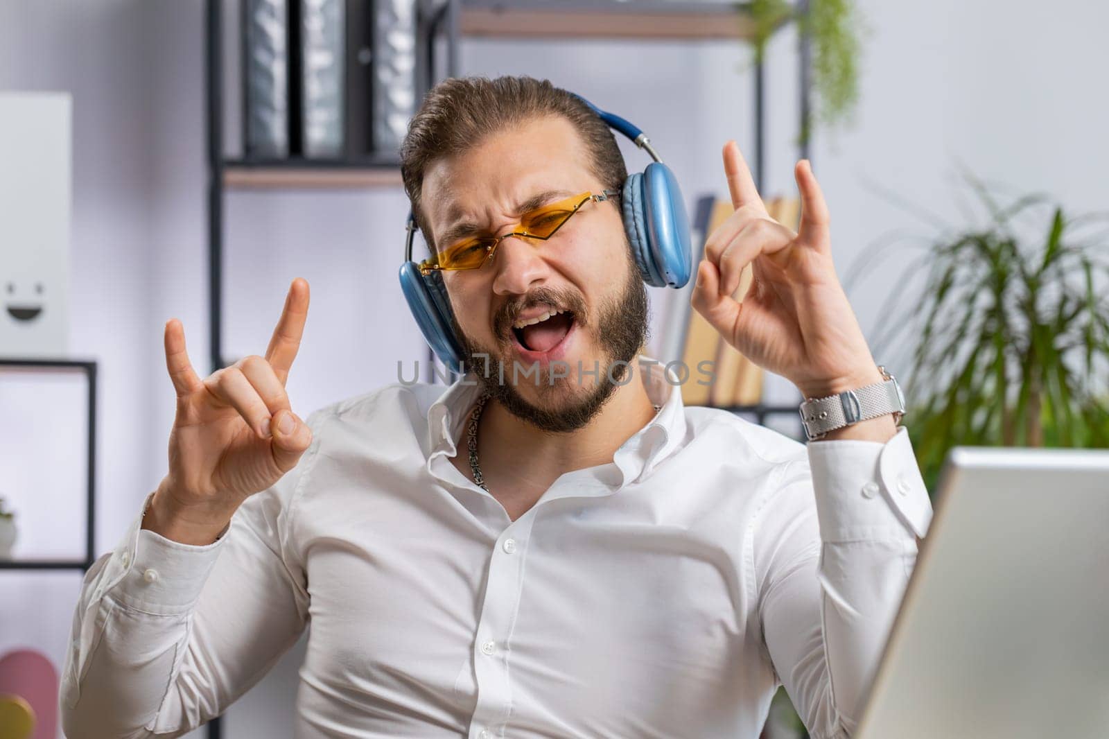Happy relaxed overjoyed middle eastern businessman working on laptop computer at office wearing headphones listening favorite energetic disco music and dancing. Freelancer man relaxing, taking a break