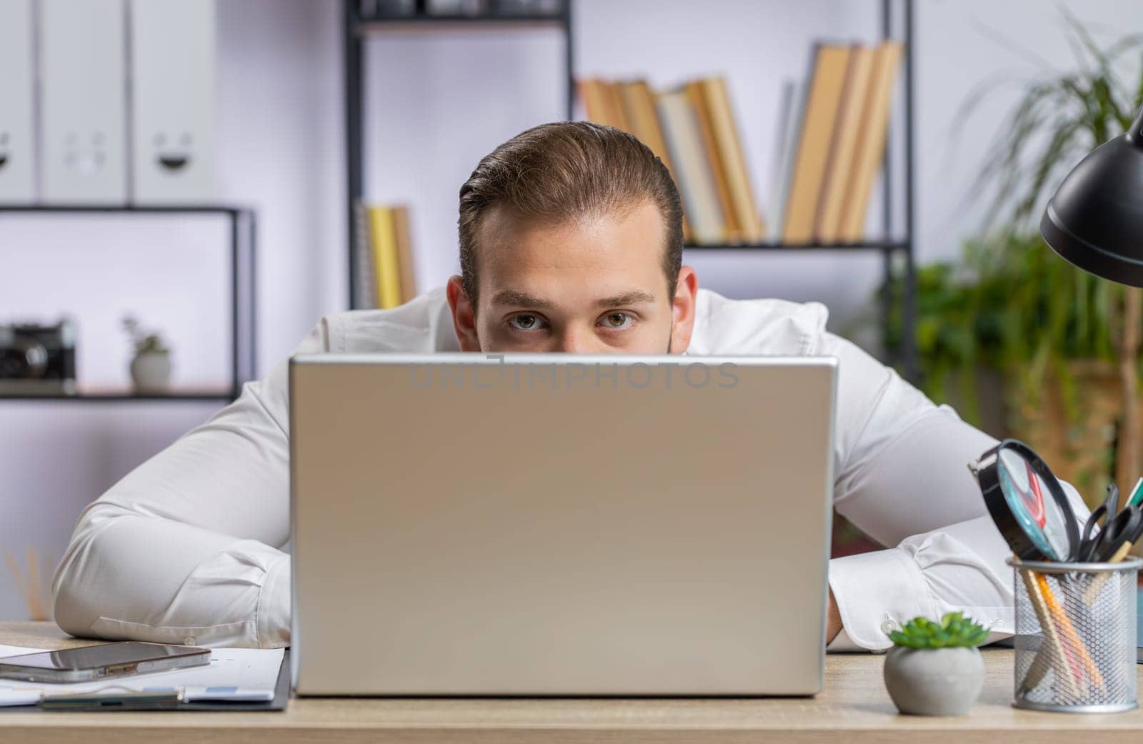 Lebanese confident businessman hiding behind laptop computer, looking at camera, spying his colleagues working, peeping. Professional freelancer man looking from behind computer with cunning eyes