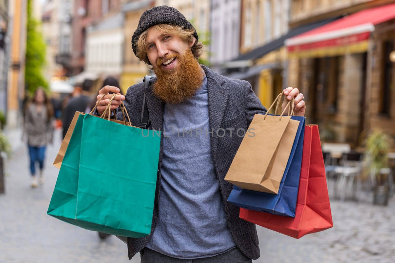 Happy smiling stylish beardedman shopaholic consumer after shopping sale with full bags with gifts outdoors. Redhead young adult guy tourist traveler walking in urban city street road. Town lifestyles