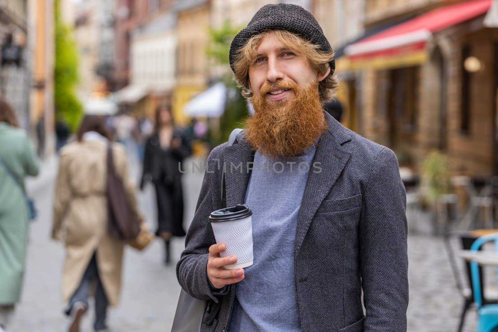 Joyful young man enjoying morning coffee or tea hot drink outdoors. Relaxing, taking a break. Redhead bearded guy walking in urban city center street, drinking coffee to go. Town lifestyles outside