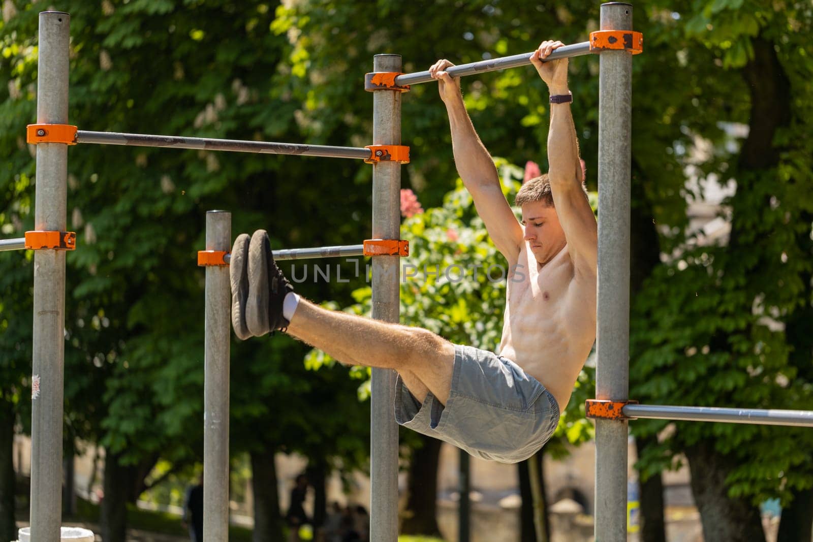 Athletic topless muscular man training abs, doing abdominal crunches, pull-ups exercises on horizontal bar. Young shirtless guy on playground. Sports health fitness routine workout strength gymnastics