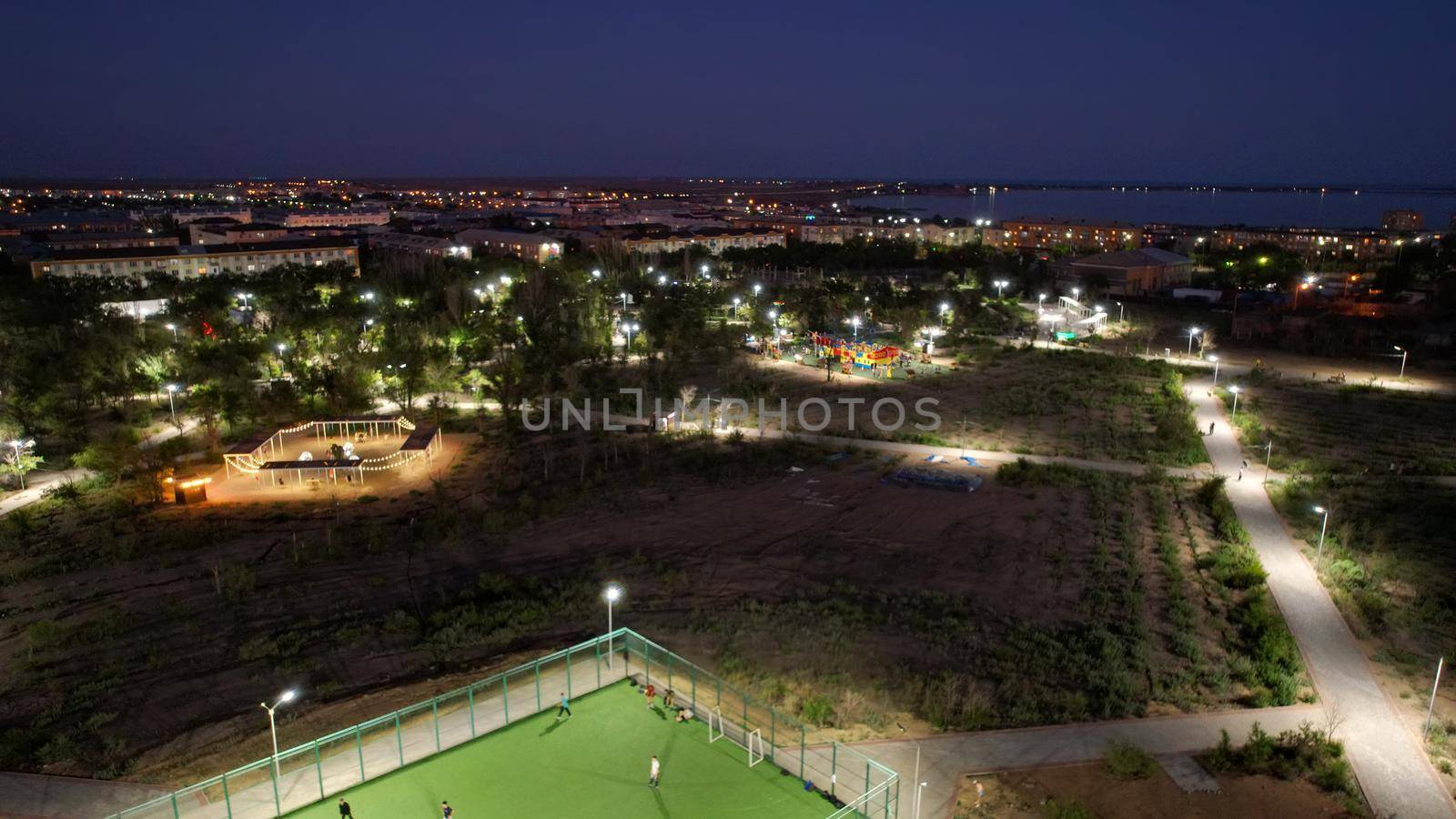 A group of people are playing football and basketball on the playground. The playground is located in the park. Lanterns are lit, people are resting and having fun. Sunset over the city. Kazakhstan