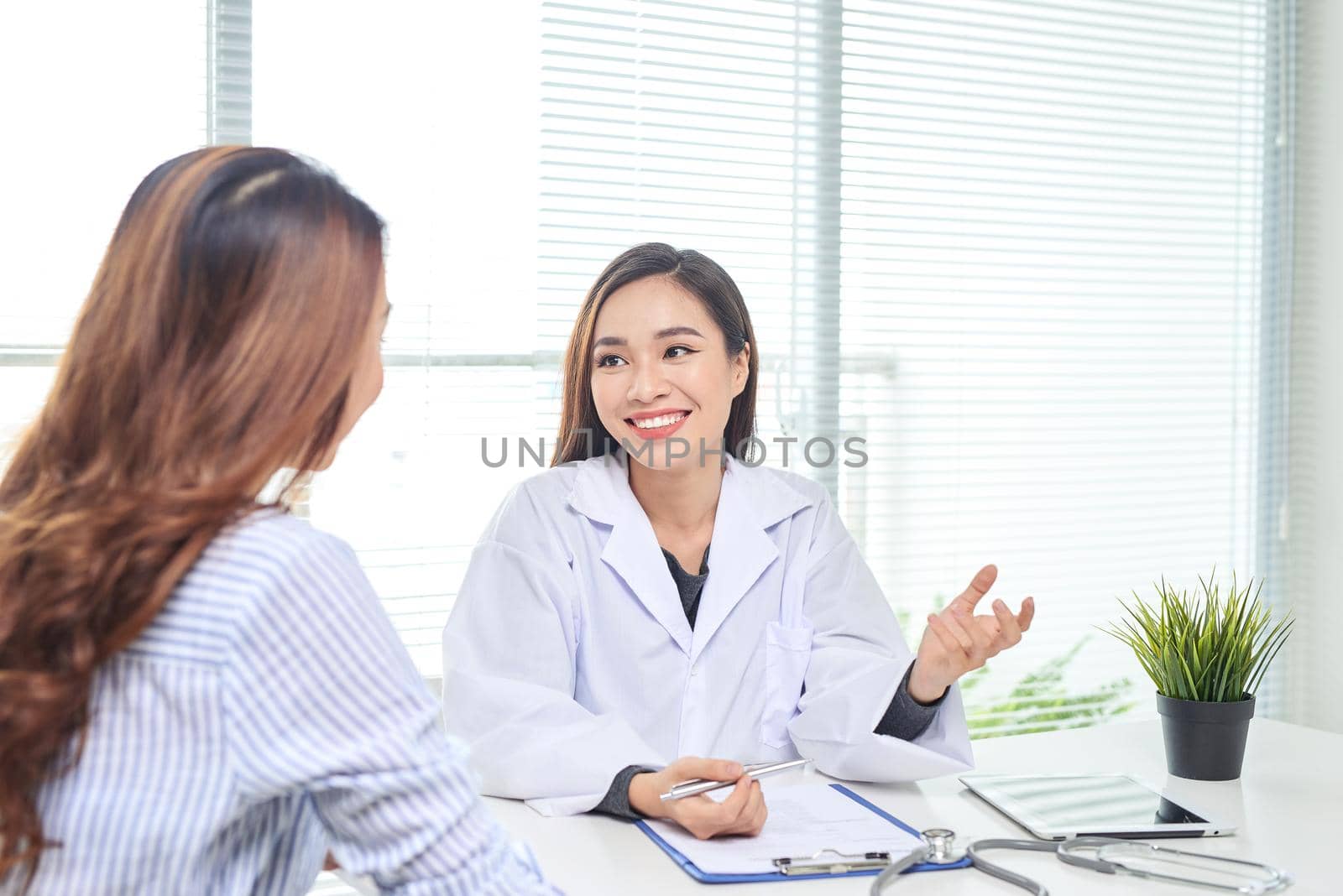 Female doctor talks to female patient in hospital office while writing on the patients health record on the table. Healthcare and medical service. 