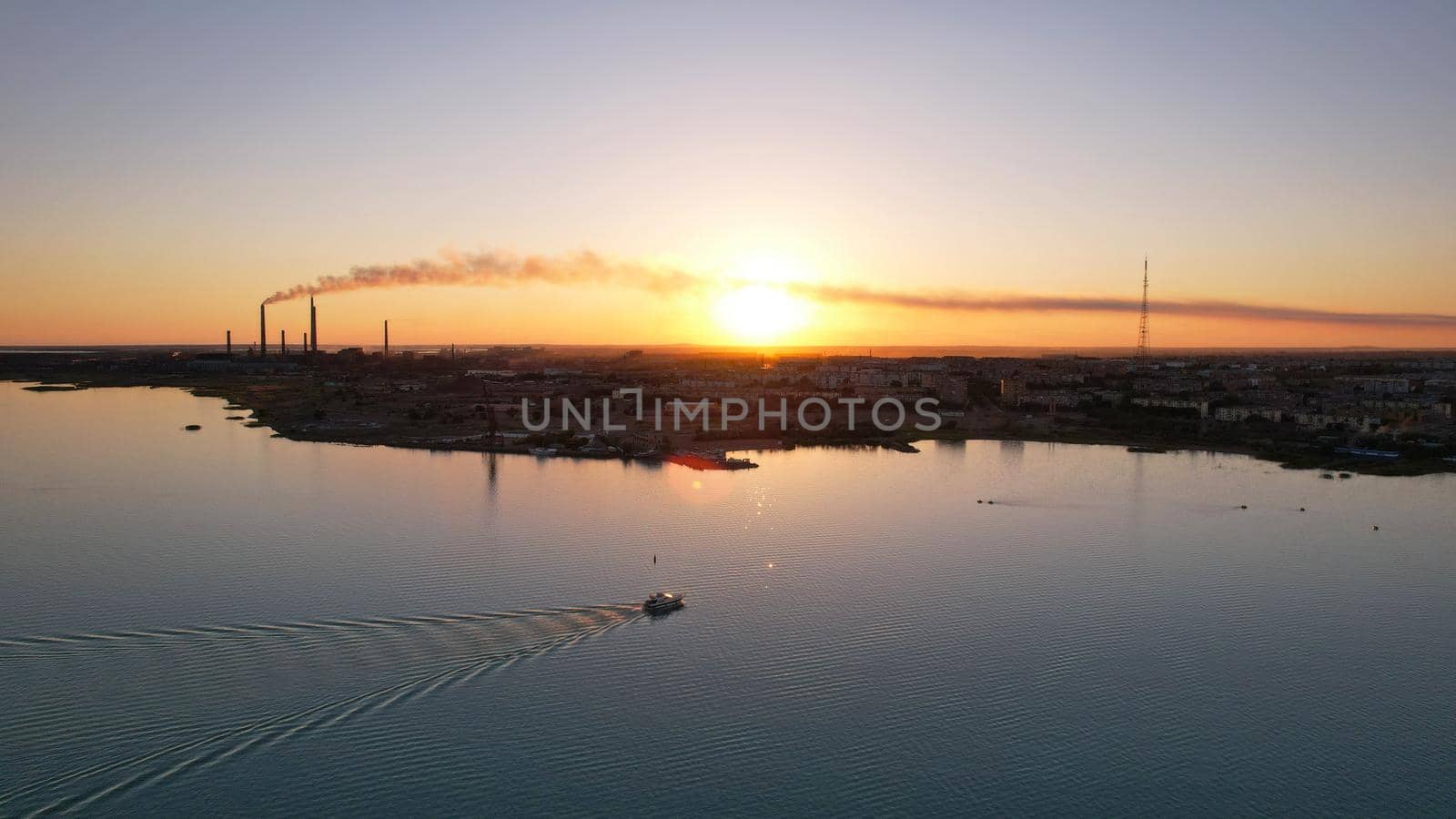 The boat sails in the bay at sunset near the city. In the distance, you can see the pipes of the plant and the orange sun. Ecology of Lake Balkhash. Catamarans float, city lights are lit. Kazakhstan