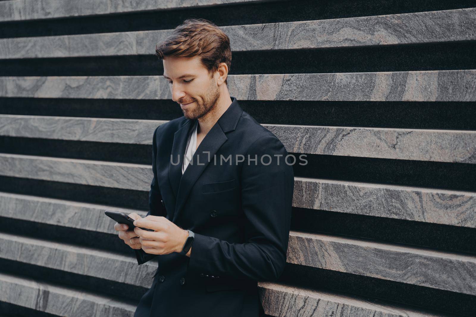 Pensive young office worker in formal stylish suit standing outdoor holding mobile phone in hands by vkstock