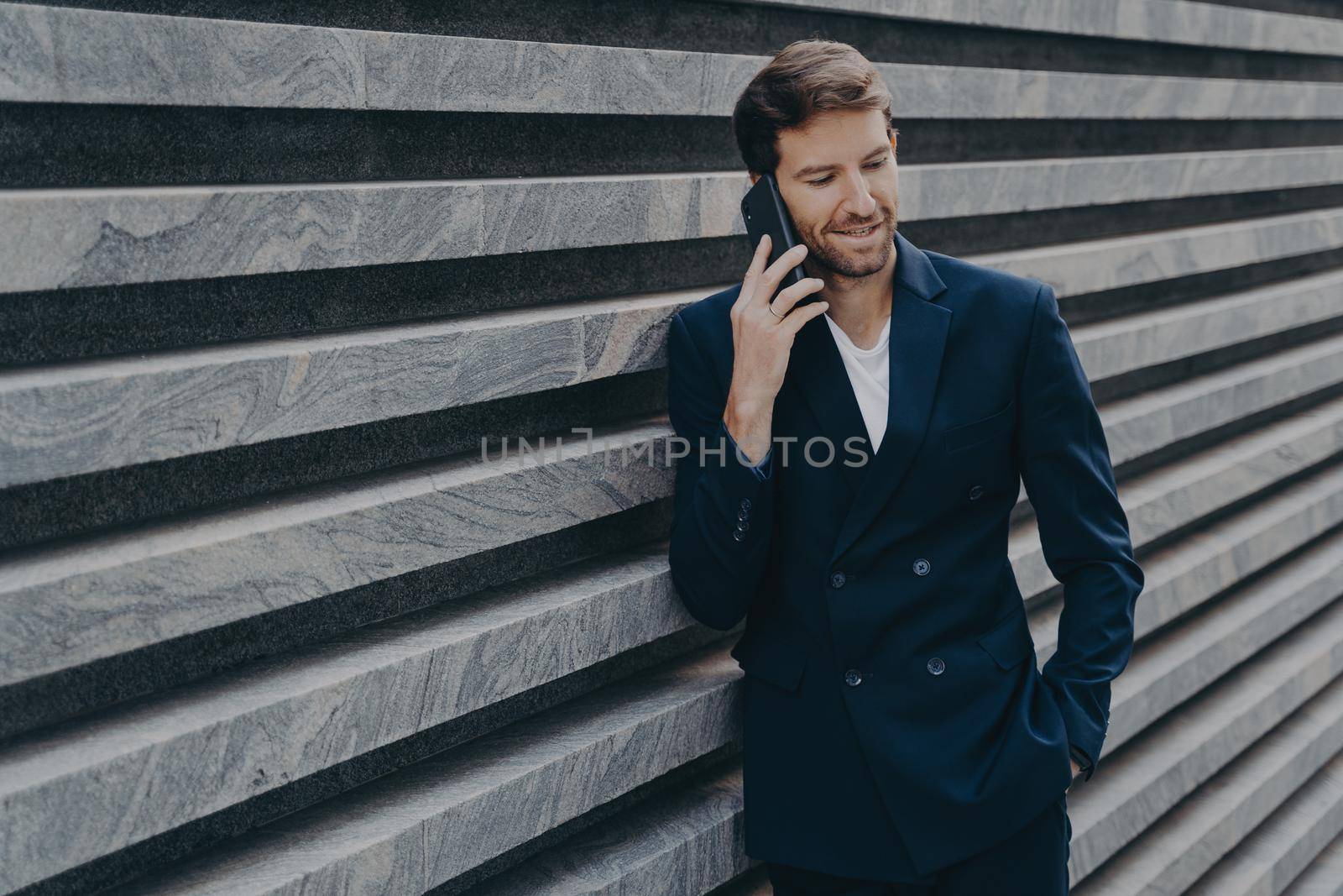 Successful male lawyer makes consultation with partner keeps hand in pocket of black suit arranges meeting via smartphone poses near marble wall copy space. People business and technology concept