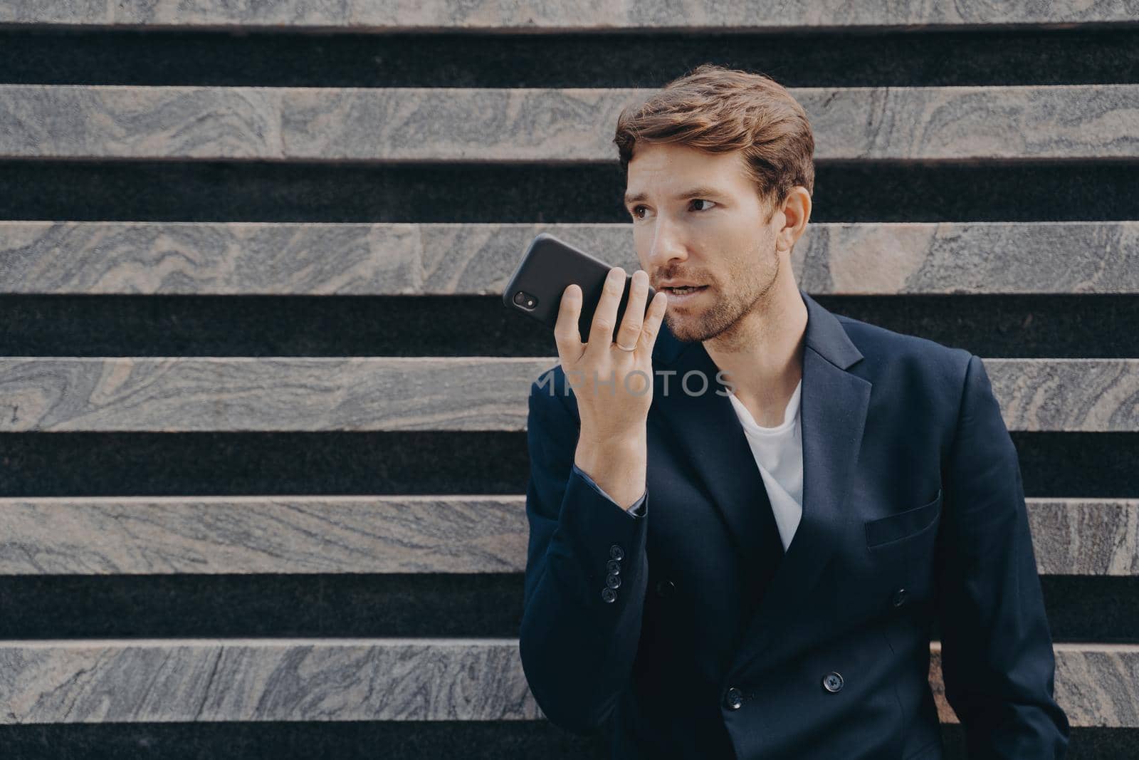 Horizontal shot of male executive worker dressed formally holds smartphone near mouth talks on speakerphone dictates voice message uses online translator app poses near marble wall copy space