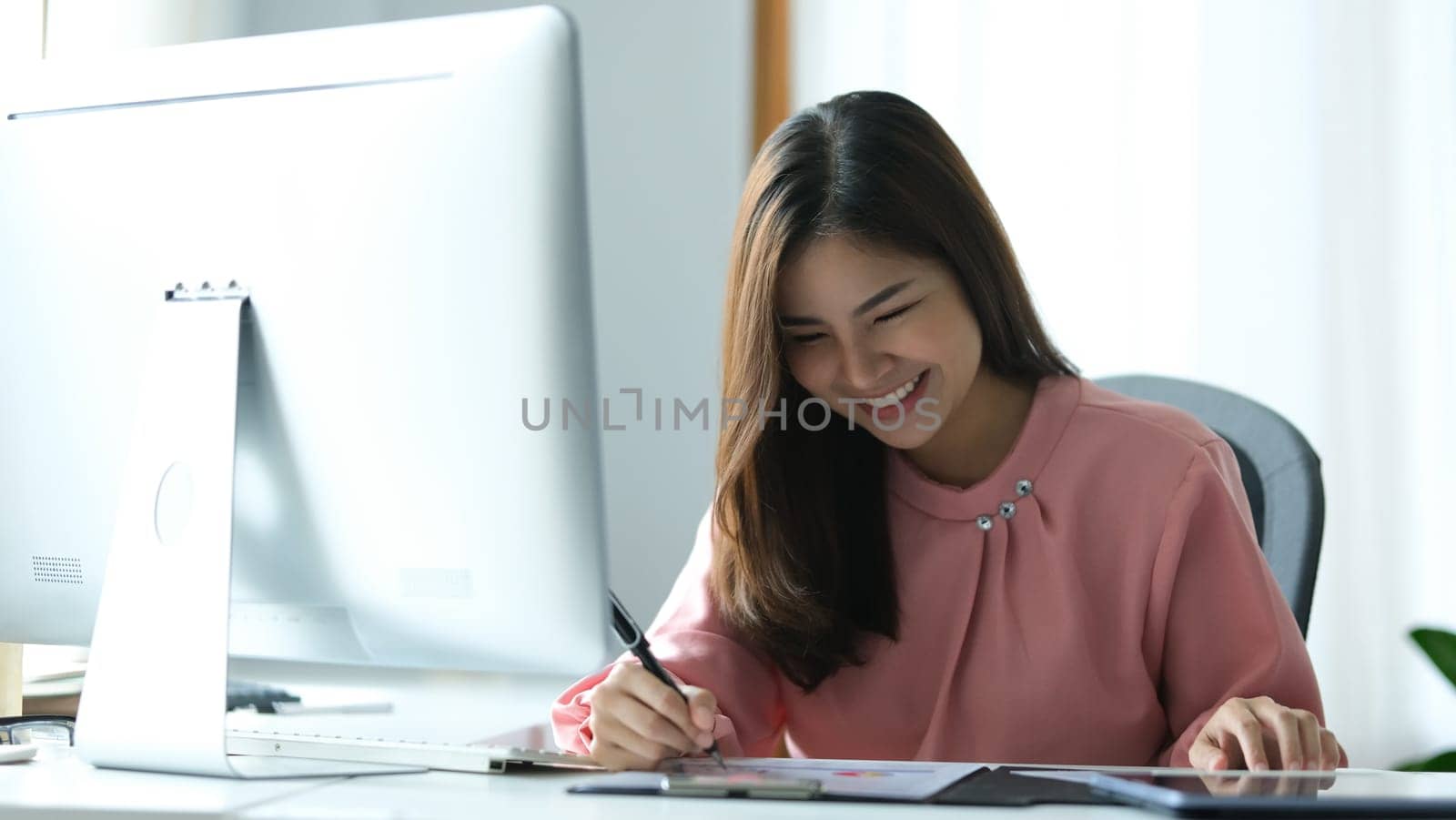 Cheerful Asian woman entrepreneur watching online webinar on computer screen and making importance notes.